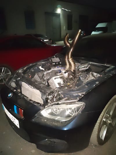 The image shows a dark-colored BMW with its hood open, revealing a heavily modified engine with prominent custom exhaust pipes. The car is parked on a paved surface, possibly a street or parking lot, at night. In the background, there are buildings with windows and doorways, and a white van. The scene appears to be in Krtsanisi, Georgia, a location known for its historical significance and rural atmosphere, although the urban elements in the background suggest a more developed area within or near the municipality. The lighting suggests artificial light sources are present. There are no people visible in the image.