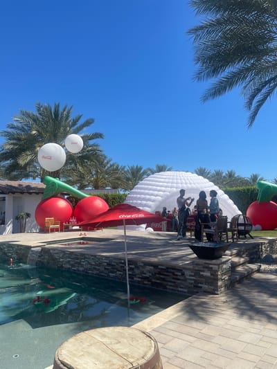 Here's a detailed description of the scene:

**Scene Overview:** The image captures an outdoor event or gathering, possibly a promotional event, on a bright sunny day in Thermal, United States.

**Environment:**  Palm trees line the area, indicating a warm climate. There is a pool present, with some possible decorations within it. The ground appears to be a combination of stone pavers and grassy areas.

**Objects:**
*   **Large Inflatables:** Several large inflatable structures dominate the scene. There is a large white dome-shaped inflatable, which might be a tent or display area. There are also two large inflatable cherries, a Coca-Cola umbrella, and Coca-Cola balloons.
*   **Furniture:** There are chairs near the white dome and Coca-Cola umbrella.
*   **Branding:** Coca-Cola branding is prominent throughout the scene, displayed on balloons and an umbrella.
*   **Wine Barrel:** A wooden wine barrel is present near the pool.

**People:**
*   There are a small number of people gathered near the white inflatable dome. They appear to be interacting with each other.
