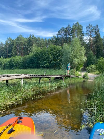The image depicts a serene natural scene in Yal'chik, Russia. In the foreground, the edge of an orange and yellow paddleboard is visible in the shallow, clear water. The water reflects the sky and surrounding trees, showing a sandy bottom in places. A narrow, weathered wooden bridge crosses the water, extending from the lush green grassy bank on the left to a sandy path on the right.
A person wearing a blue and green patterned t-shirt and shorts stands on the bridge, looking down at the water. They are wearing a light-colored bandana on their head. Tall green reeds and grasses line the edges of the water and the bridge. In the background, a dense forest of tall pine and deciduous trees forms a natural, verdant backdrop under a bright blue sky with wispy white clouds. The overall atmosphere is tranquil and suggests a warm, sunny day.
