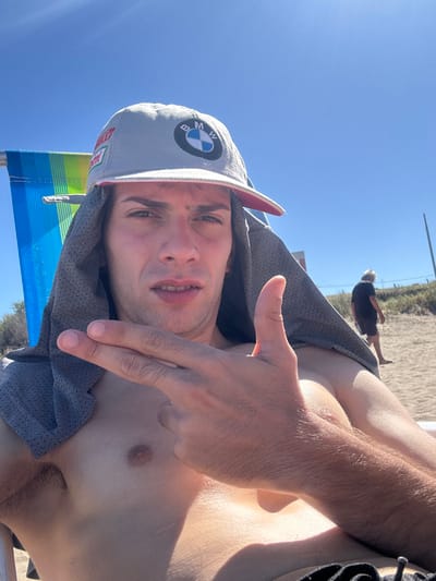 A shirtless man in a BMW hat and gray shirt reclines on a beach chair. He points a finger gun at the camera. Bright sunlight illuminates the scene. In the background, sand is visible, along with a person walking in the distance and a pole. The man is fair-skinned with short hair and facial stubble. The overall composition is casual and candid.