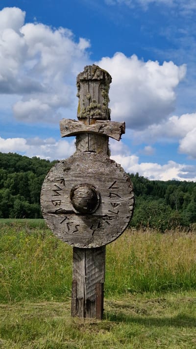 An outdoor, vertical, medium shot captures a weathered, wooden artifact resembling a Viking sword and shield, positioned centrally in a grassy field under a bright, cloudy blue sky. The artifact is constructed from rough, aged wood, showing signs of significant wear and tear, with moss and lichen clinging to its surfaces. The "sword" part is at the top, made of two pieces of wood forming a crossbar, attached to a vertical post that extends downwards. Below the sword is a round "shield" with a central circular protrusion. Carved into the surface of the shield are symbols that appear to be runes. The artifact is planted in the ground, with its base supported by two vertical metal rods. The background consists of a dense, green forest bordering a grassy hillside. The foreground is dominated by tall, dry grass, with some lighter green vegetation scattered throughout. The image is a close-up of the artifact, emphasizing its rustic texture and historical appearance. This artifact is located in Embūte, Latvia.