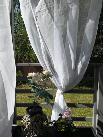 The image depicts a close-up view of a white sheer curtain blowing gently in the wind on a balcony. Through the translucent fabric, a glimpse of green foliage and a brown brick wall is visible, suggesting an outdoor setting. In the foreground, two potted plants are arranged on the balcony. The plant on the left features delicate white and pink artificial roses with green leaves, housed in a dark pot that is placed inside a woven wicker basket. The plant on the right displays small pink flowers and green foliage, also in a dark pot. A string of dark-colored fairy lights is draped across the grey railing of the balcony, partially obscured by the curtain. The overall scene suggests a bright, sunny day, possibly in a residential area of Алешкино Дп, Russia, with the natural light creating soft shadows on the curtain and railing. There are no people or animals present in the image.