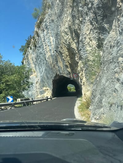 The image shows a road leading into a tunnel carved into a rocky mountainside. The tunnel entrance is dark, with daylight visible at the far end. There are two circular signs on the wall next to the tunnel entrance. One sign has a red circle with a number "3,6m" inside, indicating a height restriction of 3.6 meters. Another sign below it appears to have downward-pointing arrows. To the left of the road, a blue and white diamond-shaped road sign with a white arrow pointing to the right is visible. A metal guardrail runs along the edge of the road. The mountainside is rugged and textured, with patches of vegetation growing on its surface. The sky is clear and blue, suggesting a sunny day. The scene appears to be captured from the perspective of someone inside a vehicle, as the dashboard is visible in the foreground. This scene is located on the road D436 in the town of Septmoncel les Molunes, France, specifically at the entrance of the tunnel de Mortes-Rivières.