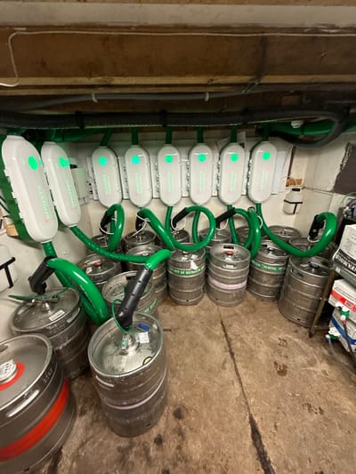 The image displays a commercial beverage cellar or storeroom. Nine white, rectangular Heineken SmartDispense units are mounted in a horizontal row on a white panel attached to a textured wall. Each unit features an illuminated green indicator light and the "HEINEKEN SmartDispense" logo. From each unit, a thick, green insulated hose descends, connecting via a black coupler to the top of an individual stainless steel beer keg. Approximately fifteen to sixteen stainless steel kegs, some branded "PROPERTY OF HEINEKEN," are arranged on a concrete floor beneath the dispensing system. Overhead, various utility pipes and cables, including green insulated lines, are visible against a low ceiling with exposed wooden joists. The concrete floor exhibits stains and appears damp in sections. On the far right, a small white plastic container is mounted on the wall, and the edge of a stack of cardboard boxes is partially visible. The scene is consistent with a commercial establishment in Sigglesthorne, United Kingdom, but no specific street or venue identifiers are present.