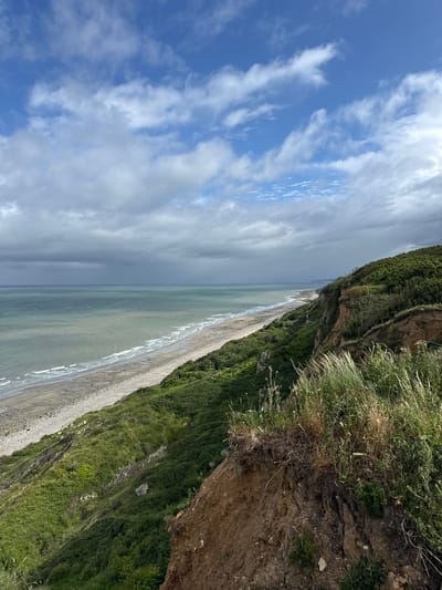 The image displays a panoramic coastal landscape in Octeville-sur-Mer, France, observed from an elevated perspective. The upper sky features patches of clear blue with scattered white cumulus and cirrus clouds, while the lower sky over the sea transitions to a uniform, darker grey overcast. The sea exhibits variations of greenish-grey and light blue, with discernible white-capped waves breaking along the extensive shoreline. The beach below consists of a mix of light-colored sand and grey pebbles/shingle, stretching horizontally across the frame into the distant background.

In the immediate foreground, a steep, eroded earth cliff face is visible, composed of reddish-brown soil with exposed root systems and sparse, dry grasses. This cliff edge descends into a densely vegetated slope covered in lush, dark green shrubs, grasses, and low-lying plants. The green-covered slopes extend along the coastline, creating a natural barrier between the land and the beach. Further along the coast, distant white cliffs are faintly visible under the grey sky, receding towards the horizon. No people, discernible man-made structures, or specific actions are present within the observable scene.