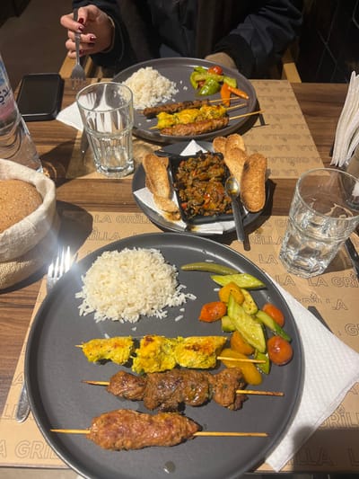 The image shows a table setting with two plates of food, each containing rice, grilled skewers, and cooked vegetables. One plate also has a side dish of what appears to be ratatouille with bread. There are glasses of water, utensils, a phone, and a wrapped loaf of bread on the table. The tablecloth or placemat has a repeating "LA GRILLA" logo. The overall scene suggests a meal at a restaurant.