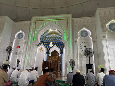 The image shows the interior of a mosque in Pasar Paloh, Indonesia, during prayers. A group of men, mostly wearing white skullcaps, are seated on a green carpet. Some are looking forward, while others are interacting with each other. The background features an elaborately decorated Mihrab with blue and gold patterns and green lighting. A wooden Minbar sits in front of the Mihrab. On the side walls, there are clock-like decorations and electric fans. A digital clock displays the time above the doorway on the left wall.