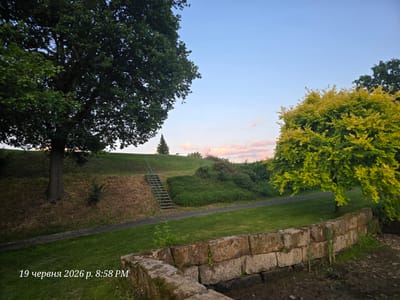 The image depicts a park-like setting at dusk. In the foreground, a rough-hewn stone wall lines the edge of a body of water. Beyond the wall, a lush green lawn stretches towards a pathway. To the left, a large, mature tree with dark green foliage stands prominently. Further back, a grassy embankment rises, with a metal staircase leading up its side. The embankment is partially covered in dry grass and dotted with bushes. In the distance, a tall, slender evergreen tree is visible on the crest of the hill. On the right side of the image, a tree with vibrant, light green foliage is illuminated by the setting sun. The sky above is a clear blue, transitioning to soft pink and orange hues near the horizon, indicative of sunset. The timestamp "19 червня 2026 р. 8:58 PM" is overlaid on the bottom left of the image. This scene is located in the city of Bannewitz, Germany.