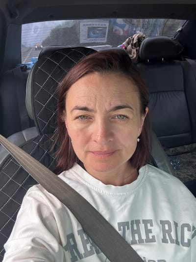 A woman is pictured in the front passenger seat of a car, wearing a white t-shirt with gray lettering. A seatbelt is diagonally across her chest and shoulder. She has medium-length dark auburn hair, green eyes, and appears to be smiling softly. A portion of a black, quilted car seat cover is visible behind her. In the background, through the car window, there are some blurred objects and what looks like a poster or advertisement. The image is taken from a high angle, looking down at the woman. The lettering on her shirt is partially visible and appears to read "THE RIC".