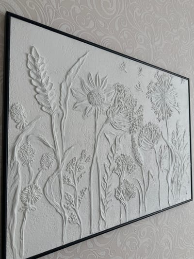 The image shows a textured white bas-relief artwork of various wildflowers, including sunflowers, dandelions, and other delicate blooms and foliage, set within a black frame. The artwork is displayed on a wall with a light-colored, embossed wallpaper featuring a swirling floral pattern. The lighting casts subtle shadows, highlighting the three-dimensional quality of the floral elements. The scene is indoors and does not provide any information about a specific location in Malye Ust'ya, Russia, or any human presence or actions.