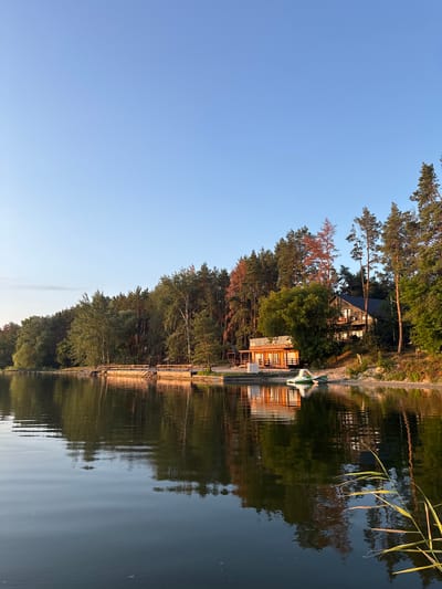 A serene lakefront scene unfolds under a clear blue sky, with the late afternoon sun casting a warm glow on the landscape. The water, calm and reflective, mirrors the lush green trees that line the opposite shore, interrupted by a wooden pier extending into the water. Further along the shore, a multi-story wooden building with a dark roof and visible windows is nestled among the trees. In front of this building, a smaller, single-story wooden structure with an open-air design and numerous tables and chairs is also visible. Two small boats, one green and white and the other green, rest on the sandy shore near the water's edge. The foreground features tall reeds growing out of the water, adding a touch of natural wildness to the scene. This peaceful setting is located in the city of Lyman, Ukraine.