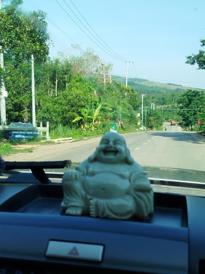 A medium shot taken from inside a vehicle, focuses on a small, light green, seated Buddha statue placed on the dashboard. The statue is smiling, and its features are clearly visible. Beyond the dashboard, the view through the windshield reveals a paved road winding into the distance. The roadside is lined with lush green vegetation, including trees and banana plants. Power lines stretch across the sky. In the distance, rolling hills can be seen under a clear, bright sky. A vehicle is driving in the distance. The location is Pờ Y, Vietnam.