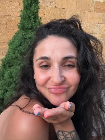 A woman with dark, wavy hair is taking a selfie in front of a brick wall and a green, conical bush. Her lips are pursed as she blows a kiss towards the camera, her right hand extended with painted fingernails. She has freckles across her nose and cheeks and a tattoo on her left forearm. The setting appears to be outdoors, possibly in a residential area.