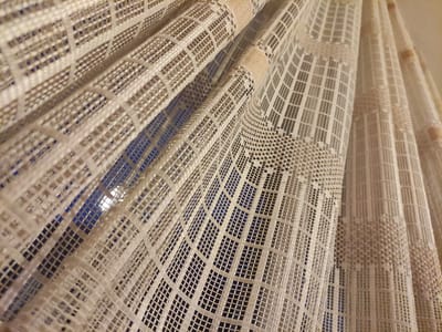 The image displays a close-up view of a white lace curtain with a checkered pattern. The curtain is layered and draped, with folds and creases creating a sense of depth. The fabric is semi-transparent, allowing a glimpse of a blue background, likely a window or wall, with some light sources visible through the mesh. The lighting in the scene is soft, highlighting the texture and intricate details of the curtain. There are no people or specific objects visible beyond the curtain itself. The scene appears to be indoors, possibly in a residential setting. The provided location of Krushev Dol, Bulgaria, is noted, but there are no visual cues within the image to confirm or deny this specific location; it simply depicts a curtain.