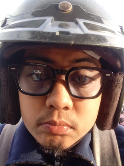 A close-up, head-on, outdoor selfie shows a person wearing a dark motorcycle helmet and black-rimmed glasses. The person's face is lit by sunlight from the right, casting shadows to the left. Their eyes are visible through the glasses, with the left eye appearing slightly more in focus. A thin mustache and goatee are visible on the person's upper lip and chin. The background is out of focus, but appears to be an outdoor environment, possibly in the city of Tocina, Indonesia. The person is wearing a dark blue jacket with a gray strap over their right shoulder. A hint of fabric, possibly part of a backpack, is visible to the left of the person's head. The helmet has a gold emblem on the top front and appears to be well-worn.