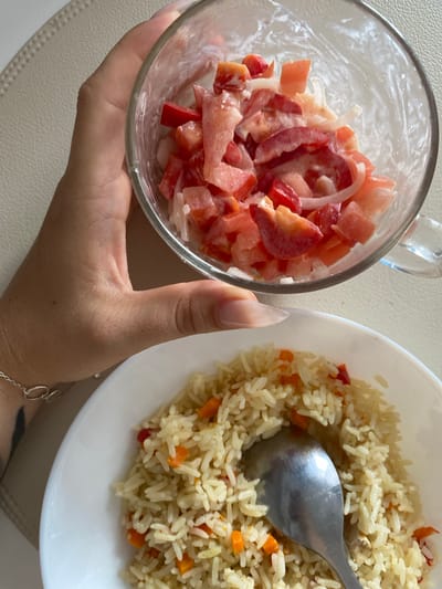 A close-up, eye-level shot shows a person's hand holding a glass bowl filled with chopped tomatoes and onions mixed with a creamy dressing. Below the bowl, a white plate contains a portion of rice pilaf with diced carrots and peppers, with a spoon resting in the dish. The background is a light-colored, textured surface, possibly a placemat or table. The image is shot in Ostrovki, Russia, implying an indoor setting given the lighting and composition.
