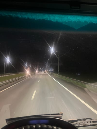 A wide-angle, low-light, forward-facing perspective captures a highway at night, viewed from inside a vehicle. The road ahead is illuminated by numerous streetlights, creating bright, star-like flares due to the camera's exposure. The highway has visible white lane markings and a grassy median with a guardrail on the left. The right side of the road is bordered by darkness, suggesting a rural or undeveloped area. In the distance, headlights of other vehicles are visible. The upper portion of the image shows the car's windshield, with a hint of a blueish tint at the very top edge, possibly from the car's interior or a tinted strip on the windshield. The foreground shows the top of a dashboard and windshield wipers. The scene is devoid of people and any specific identifiable landmarks that would definitively place it in Vladimirovka, Russia, or any other city.