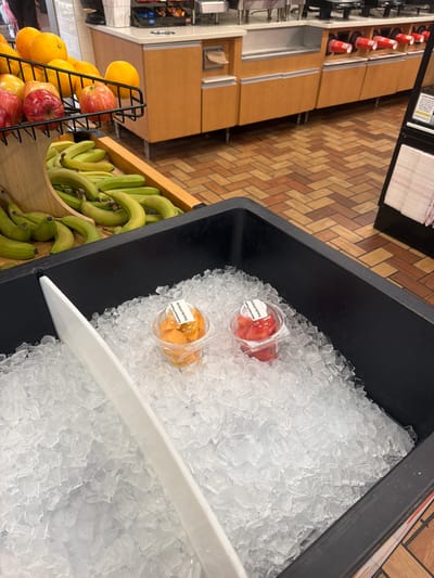 The image shows a close-up view of a black plastic bin filled with crushed ice, positioned at a service counter within what appears to be a convenience store or gas station in Raphine, Virginia. Two small, clear plastic cups containing diced fruit are visible, embedded in the ice. The cup on the left contains orange-colored fruit, likely cantaloupe, and the cup on the right contains red fruit, likely watermelon. Both cups have white labels that are partially legible, one indicating "Enjoy by: 07/17/2023". A white plastic scoop or divider is also present in the ice. In the background, a display rack with oranges, apples, and bananas can be seen. Further in the background, a counter with beverage dispensers and red paper cups is visible, with wooden cabinets and patterned flooring completing the scene. There are no people visible in the frame.