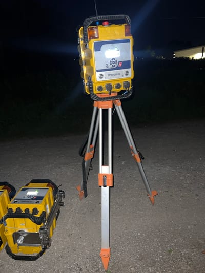 The image depicts two yellow Zollner ZPW126-10 devices, likely used for surveying or measurement, set up on a gravel surface at night. One device is mounted on an orange and silver tripod, with its screen illuminated. The other device is partially visible in the foreground, also appearing to be set up for use. The background shows dark vegetation and a lit building in the distance, suggesting an outdoor location in a semi-rural or industrial area. The context provided by the user places this scene in Bamberg, Germany, although this cannot be visually confirmed from the image alone. There are no people visible in the image.