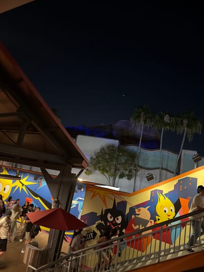 The image shows a nighttime scene at a location in Anaheim, USA. Tall palm trees are visible in the mid-ground, illuminated with blue light. Cartoon character themed building with a yellow character with hair that is yellow. The foreground reveals a partially visible metal and wood structure. An open red umbrella is also in the foreground. People are walking around in the background, including some that are standing under the umbrella. A railing and stairs are running along the side. The sky is dark, with a small crescent moon visible in the upper left.