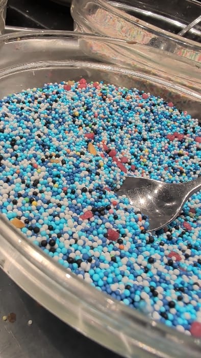 A clear serving dish, likely glass or plastic, is depicted filled with a dense collection of spherical confectionery sprinkles. The sprinkles primarily consist of various shades of blue, white, and black, intermixed with smaller quantities of orange, yellow, and red spheres. Several irregularly shaped red and yellow confectionery pieces are also present. A silver-colored metal spoon is partially submerged in the sprinkles on the right side of the dish. The dish rests on a dark, reflective surface. A portion of another clear container is visible in the blurred background. This scene is located in Horst, Netherlands.