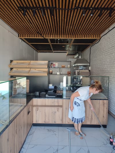 The image shows a woman in a white t-shirt and a checkered apron sweeping the floor of a commercial space. The space appears to be a small shop or bakery, with a counter and display cases visible. The walls are white brick, and the ceiling has a wooden slatted design with track lighting. There are shelves with wooden trays mounted on one wall, and a commercial ventilation hood above the counter. A cane stands beside the woman. In the foreground, there are bottles of liquid, possibly beverages, near the floor. The floor itself is tiled with a marble-like pattern. The overall impression is one of a business undergoing cleaning or preparation for opening.