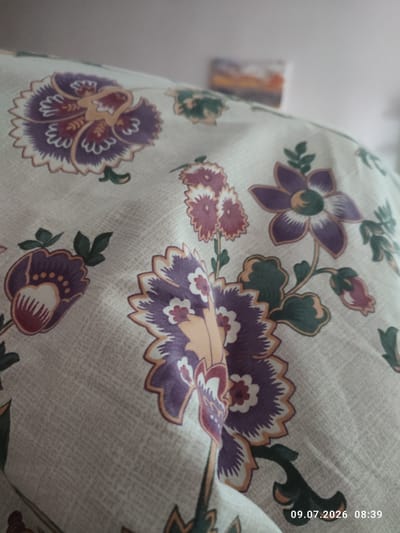 This image depicts a close-up view of a fabric with a floral pattern. The pattern features stylized flowers in shades of purple, pink, yellow, and green, set against a light-colored background with a subtle texture. The fabric appears to be draped or folded, creating soft shadows and highlights that emphasize the curves and folds. In the upper background, a blurry image of a landscape painting or photograph can be seen. A timestamp in the bottom right corner reads "09.07.2026 08:39". The image does not provide any identifiable landmarks or elements that would allow for pinpointing a specific location within Цярэшкавіцкі сельскі Савет, Belarus.