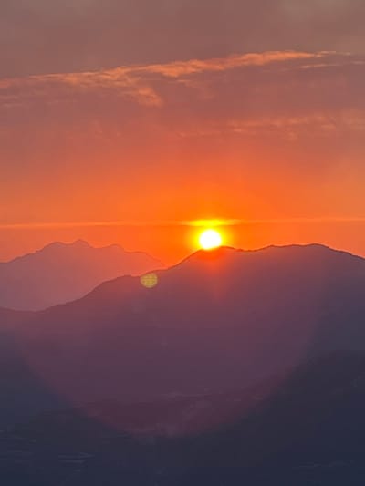 The image depicts a scenic mountain landscape at sunset. The sun, a bright orb, is partially visible, casting a warm orange glow across the sky and illuminating the peaks of several layered mountain ranges. The foreground mountains are silhouetted in shades of purple and dark blue, suggesting depth and distance. Wispy clouds are present in the upper portion of the sky, tinged with the same vibrant sunset colors. The scene is devoid of human presence. The location is San Juan de Plan, Spain, a town situated within the Pyrenees mountain range, known for its dramatic natural beauty.