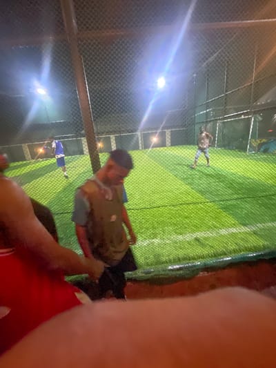 The image depicts a nighttime scene at a soccer field, likely in Malete, Nigeria, illuminated by bright floodlights. In the foreground, out of focus, is a person wearing a red garment, possibly shorts. To the left, another person's arm, also out of focus, is visible.

In the mid-ground, a young man is standing and looking downwards, wearing a light brown vest over a blue t-shirt and dark pants. Behind him and to the left, another individual in a blue jersey and shorts is walking away from the camera, on the green artificial turf of the soccer field. Further in the background, to the right, a person in a patterned shirt and light-colored pants stands with their hands on their hips.

The soccer field itself is well-lit and appears to be enclosed by a mesh fence. The ground is covered with artificial green turf, marked with white lines. The overall atmosphere suggests a casual sporting or recreational activity taking place after dark.
