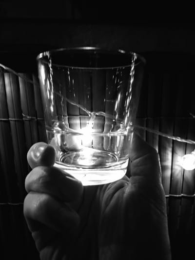 A close-up, black and white, low-angle shot shows a hand holding a clear glass filled with a liquid, likely water, indoors. The hand is gripping the glass from below. The glass has a string of small, round lights, possibly fairy lights, wrapped around it, and some of these lights are illuminated, casting a soft glow through the liquid. In the background, a textured, dark surface, possibly a bamboo mat or blinds, is visible, with more of the string lights scattered behind it. The lighting is dim, with a bright light source reflecting within the glass. The scene is devoid of any identifying landmarks or characteristics that would associate it with Sinzheim, Germany.