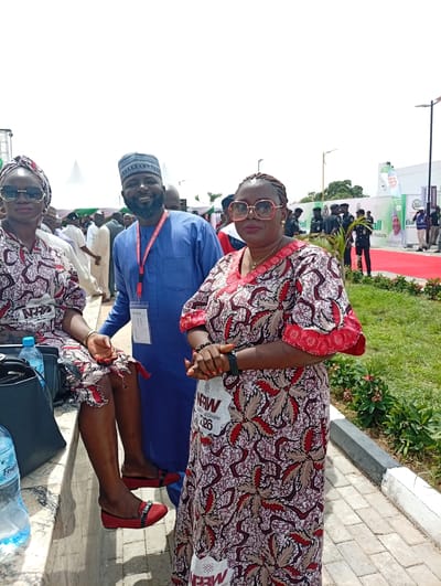 This outdoor photograph captures three individuals posing at an event in Unguwan Sanusi, Nigeria, under bright daylight. In the foreground, a man wearing a blue kaftan, a traditional patterned cap, and a beard smiles broadly. He has an ID badge around his neck which prominently features the Chinese characters "工作证" (Work Permit), alongside other text like "APE CROC CITY 200" and "NAFD."

He is flanked by two women dressed in matching patterned outfits featuring red, brown, and white designs. To his left, a woman is seated on a low concrete wall or bench, wearing a headwrap, dark sunglasses, and red flat shoes. A "Faya" brand water bottle and a black handbag are visible beside her. To the man's right, another woman stands, wearing red-rimmed glasses and an ID badge that reads "NRW 226 State Chapter" and "Kalimahanter."

The background indicates a large outdoor event, possibly a conference or festival. Tents and various banners are visible, with text including "Hall," "Kaduna," and "ENBRIDGE." A red carpet stretches into the distance, and uniformed personnel along with other attendees can be seen further back. The bright, clear sky suggests it is daytime. The scene is set on paved ground with patches of green foliage, contributing to a vibrant and communal atmosphere.