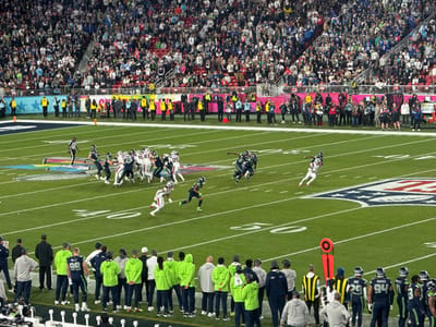 An American football game unfolds on a green field, with players in contrasting blue and white uniforms engaged in a play near the center of the field. Lines and numbers mark the field, and an orange marker stands near the sideline. Spectators fill the stadium, creating a dense backdrop of faces. Along the sideline, players and coaches in green and blue watch the game intently.