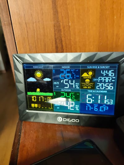 A digital weather station displays various weather-related information. The station shows the forecast for sunny and partly cloudy weather with a dandelion in the foreground. Indoor temperature is 26.1°C with 54% humidity, and outdoor temperature is 24.5°C with 42% humidity. The station also indicates sunrise at 4:46 AM and sunset at 8:56 PM for the city of PAR. The current time is 6:11 AM and the date is 17-6 CP. The device is placed on a wooden surface, possibly a table or shelf, in front of a wooden cabinet. No people are visible in the image. The location is identified as Bashkia Durrës, Albania.