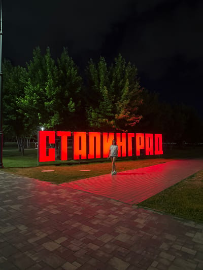 An outdoor, nighttime shot shows a large, illuminated red sign that reads "СТАЛИНГРАД" (Stalingrad). The sign is composed of large, blocky, capital letters, each individually lit from within and mounted on red supports. A young woman wearing a white t-shirt and dark shorts stands to the left of the sign, facing the camera. She is wearing white sneakers. The sign is situated in a park-like setting with green grass, mature trees, and a paved walkway. The walkway, in the foreground and extending to the right of the sign, is paved with interlocking stones, some of which are illuminated by a red glow originating from the sign and possibly nearby lighting. The background is dark, suggesting it is late at night, with the dark shapes of trees and what appear to be streetlights visible. The setting is the city of Volgograd, formerly Stalingrad, Russia.