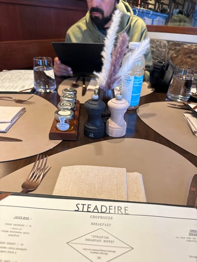 The image depicts a restaurant breakfast scene at Steadfire Chophouse, located in Teton Village, United States of America.
In the foreground, a dark wooden table is set with light brown, curved placemats. A black menu, titled "STEADFIRE CHOPHOUSE BREAKFAST," is visible in the lower center. To the left, a copper-colored four-tined fork rests on a placemat beside a folded beige napkin. On a central placemat, a small dark vase holds a floral arrangement of white and light purple dried pampas grass plumes. To its immediate right, a black Peugeot pepper grinder and a light wood Peugeot salt grinder stand. Behind these, a plastic bottle of "zero sugar" beverage is visible.
Further left on the table, a wooden rectangular tray holds five small lidded glass jars, one of which is labeled "PURE HONEY." A clear glass filled with water is positioned to the left of the tray.
Seated behind the table, partially visible from the chest up, is an adult male with a dark beard and mustache, wearing a green hooded sweatshirt. He is holding a black menu or tablet with both hands, appearing to read it. To his right, a second clear glass of water and a black camera bag or similar item are visible on the table. Cutlery, including a knife and spoon, are placed near his right hand. The background features dark wooden paneling and stone wall sections, indicating an indoor dining establishment.