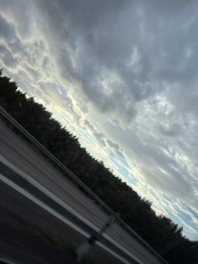 This image captures a view from a moving vehicle on a highway, likely in or near Löftaskog, Sweden. The perspective is angled, looking towards the sky and a dense line of dark green trees bordering the road. The sky is dominated by dramatic, gray and white clouds, with patches of blue sky visible between them. Sunlight is breaking through in some areas, creating highlights on the clouds. The highway itself is visible in the foreground, with dark asphalt and white lane markings. A metal guardrail runs along the side of the road. The overall impression is one of a somewhat overcast and dynamic sky, seen from a typical roadside environment.