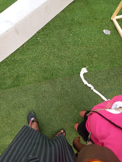 The image depicts a ground-level view of two individuals standing on green artificial turf in Ohafia, Nigeria. The individual on the left is wearing black and dark grey striped trousers and black flip-flops with red and green striped accents. The individual on the right is wearing a bright pink top, dark legwear, and light brown boots; a black strap, possibly from a bag, crosses the pink top, which also features a white patch with partially visible text, "E. Francia." The environment includes a white concrete or painted barrier running diagonally along the upper left edge and a section of a light wooden frame in the upper right. Scattered debris on the turf consists of a small grey object near the wooden frame and a crumpled white piece of material in the mid-ground. The individuals are positioned side-by-side, their feet in close proximity.