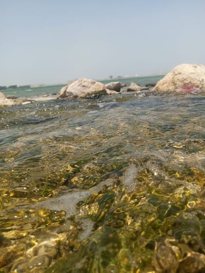 The image shows a close-up view of shallow water near a rocky shore in Al `Aziziyah, Saudi Arabia. The foreground is dominated by the water's surface, which is rippled and translucent, revealing pebbles and green algae on the seabed below. Small bubbles are visible on the water's surface. In the background, large, light-colored rocks are scattered along the shoreline. Beyond the rocks, a strip of turquoise sea and the distant city skyline are visible under a pale blue sky. The scene appears to be captured on a bright, sunny day.