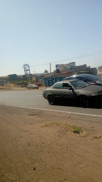 A street scene reveals a gray sedan in the foreground, its paint appearing faded. Behind it, another car is partially visible. Across the street, a light-colored hatchback is parked near a roadside establishment. Buildings and signs line the background, overshadowed by a clear sky. A leaning utility pole adds a slight disturbance to the horizon. The foreground is dominated by a dirt shoulder, with sparse vegetation.