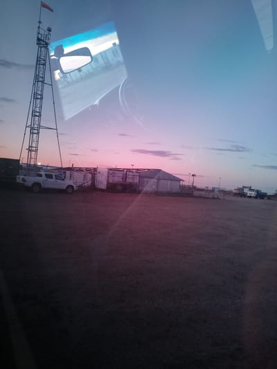 The image depicts a rural scene in the city of Zhasuat Rural District, Kazakhstan, during a sunset. The sky displays a gradient of soft blues and pinks, indicating the twilight hours. In the foreground, a vast, unpaved area stretches towards the horizon. A white pickup truck is parked near a series of modular buildings or temporary structures, some of which appear unfinished or under construction, as evidenced by exposed framing. To the left, a tall, metal tower with a windsock at its apex stands prominently. In the background, several construction vehicles, including a white truck and a yellow excavator, are visible. The reflection in the pickup truck's side mirror shows a glimpse of the vehicle's interior, including the dashboard and steering wheel, and the tower structure. The overall impression is one of an industrial or construction site settling in for the evening.