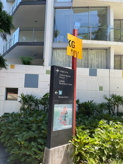 This image captures a bright, sunny daytime scene featuring a modern building facade, lush green foliage, and a prominent directional sign, likely in Kelvin Grove, Australia.
The main subject is a black, multi-panel directional sign standing in front of a white building. The sign features a yellow, arrow-like element at the top, displaying the letters "KGV". Below this, the black panel provides navigational information. An upward-pointing arrow indicates "Village Centre", "Creative Industries Precinct", "La Boite", and "Round House Theatre". A downward-pointing arrow points to "Busway Station". The sign also includes a small, colored map or diagram under a clear cover, depicting various sections of an area with a red path highlighted.
Behind the sign, the building features a white facade with varied textured panels and several large windows that reflect the sky and surrounding trees. Visible text on the building's facade includes "HORIZONS" near the top right and "VISTA" lower down. Patches of green and blue square tiles are integrated into the white wall design.
To the right of the sign and building, dense, bright green foliage with deeply lobed leaves grows, contributing to a vibrant, natural feel. More greenery is visible near the base of the building. The scene appears well-lit, suggesting clear weather conditions. No people or activities are explicitly depicted.