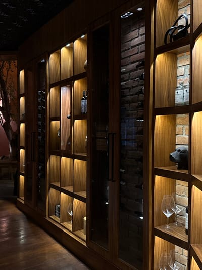 An upward shot shows a wooden shelving unit with various items. Some shelves have wine bottles, others hold wine glasses, a black cable, and a dark sculpture resembling a dog. Above these shelves is a mirrored panel showing a brick ceiling with recessed lighting. The bottom shelves are dimly lit and reveal glimpses of an outdoor scene with a tree.