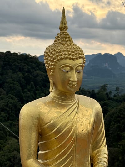 A close-up, eye-level, vertical shot features a golden Buddha statue against a backdrop of lush, green mountains under a cloudy, pale yellow sky. The statue, which is the primary subject, is centrally located and fills most of the frame. It is facing slightly to the right, with its gaze directed downwards and to the side, conveying a serene and contemplative expression. The statue is adorned with traditional robes, which are intricately folded and draped across its body. The gold surface of the statue shows some wear, with patches of lighter gold and darker undertones, suggesting age and exposure to the elements.
The background consists of dense, dark green foliage covering the rolling hills, characteristic of the tropical landscape of Ban Khao Tang, Thailand. In the distance, the hazy outlines of karst mountains rise majestically, adding depth and a sense of grandeur to the scene. The sky is overcast with streaks of pale yellow light filtering through, indicating either sunrise or sunset. A thin, dark wire is visible crossing the left side of the frame, a subtle man-made element in an otherwise natural and spiritual setting.