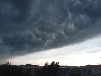 This image captures a dramatic sky over the city of Kyshtym, Russia, likely taken from an elevated vantage point. The sky is dominated by dark, ominous storm clouds that fill the upper two-thirds of the frame. These clouds have a turbulent, textured appearance, with deep shadows and lighter, swirling patterns. Below the storm clouds, a sliver of lighter sky is visible, transitioning to a hazy, pale sunset glow near the horizon.
In the foreground, the silhouettes of apartment buildings are visible, characteristic of urban Russian architecture. These buildings appear to be several stories high, with numerous windows and balconies. Dark trees, their branches appearing windswept, are scattered in front of and between the buildings, suggesting strong gusts of wind accompanying the approaching storm. The overall impression is one of an impending weather event, with the city bracing for a storm. There are no visible people or animals in the frame. The scene is devoid of any discernable actions or interactions.