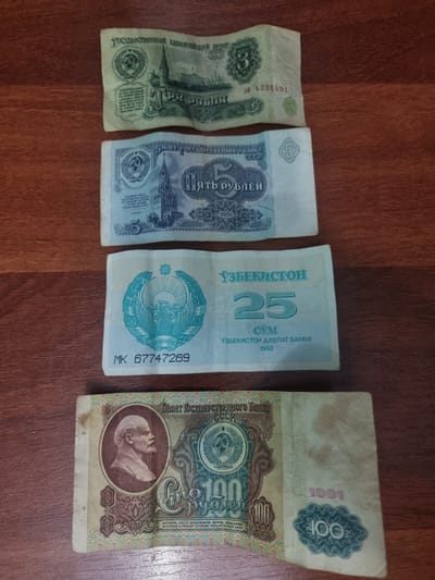 The image shows four pieces of currency laid out on a wooden surface. The currency appears to be from different countries and eras.
From left to right, the first bill is green and has text that appears to be in Cyrillic script. Visible text includes "ГОСУДАРСТВЕННЫЙ КАЗНАЧЕЙСКИЙ БИЛЕТ" and "1961". It also has a serial number "CÓ 4226491".
The second bill is light blue and features a building and Cyrillic text. Prominent text includes "ПЯТЬ РУБЛЕЙ" and "ГОСУДАРСТВЕННОГО БАЙНА".
The third bill is light blue and has text in both Cyrillic and Latin scripts. It prominently displays "25" and "CYM" in large font, along with "ЎЗБЕКИСТОН" and "ЎЗБЕКИСТОН ДАВЛАТ БАНКИ 1992". A serial number "MK 67747269" is also visible. This bill is from Uzbekistan.
The fourth and largest bill is light brown and features a portrait of a man, likely a political figure. It displays "100" in large font and has Cyrillic text including "ГОСУДАРСТВЕННОГО БАНКА СССР" and "1991". It also has text that includes "БАНКОВСКИЕ БИЛЕТЫ ОБЕСПЕЧИНАЮТСЯ ЗОЛОТОМ".
The wooden surface suggests an indoor setting, likely a table. The lighting is even, indicating no specific time of day or weather conditions are evident. The objects are the sole focus of the image.