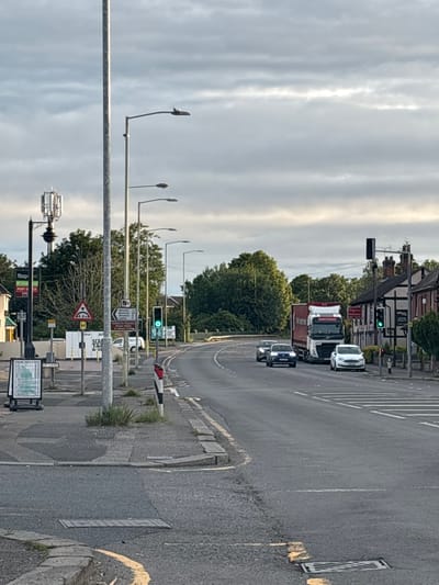 The image depicts a street scene in South Staffordshire, United Kingdom. A grey, overcast sky with textured clouds forms the backdrop. In the foreground, a two-lane road runs horizontally, with white road markings and a yellow curb. A number of streetlights are visible along the side of the road, as well as a tall pole with an antenna at the top.
Several vehicles are present: a white articulated lorry, a white Ford car, a dark blue car, and a second dark blue car further down the road. There is a traffic light that is currently green. On the right side of the road, there are buildings with Tudor-style architectural elements, and dense green trees line the background. Signs for "Whiston, Brocton, Ingestre, Stafford" and a red and white striped barrier are visible. A white pedestrian crossing is marked on the road. On the left side of the road, there is a post box, a "Co-op" sign, and a sandwich board advertising parking facilities. The pavement has some patchy grass growing through it.