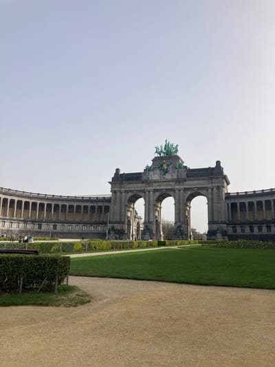 The image depicts the Cinquantenaire Arch and surrounding park in Etterbeek, Belgium. A large, grey stone triumphal arch featuring three main archways stands centrally. Perched atop the arch is a prominent bronze quadriga sculpture depicting a four-horse chariot with figures, situated above a gilded crest. Flanking the arch are two expansive, curved colonnaded buildings, also constructed of light grey stone, each featuring numerous columns and windows.
In the foreground, a wide, light brown gravel path curves towards the right. This path transitions into a section of neatly mown green grass, bordered by a dark green, low-trimmed hedge. Further back, another large expanse of green lawn extends towards the architectural structures. A few individuals are visible in the distance on the left side of the park, near the base of the curved building, appearing to be walking or standing. The sky above is clear and light blue, indicating daylight conditions with subtle shadows.
