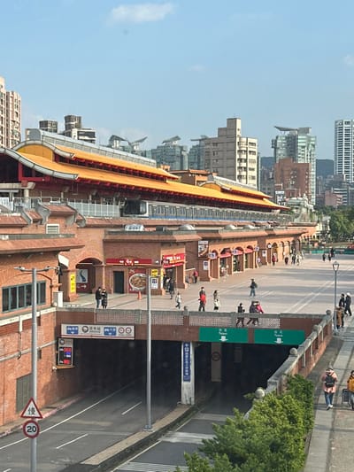 A long, traditional building with a yellow tiled roof stands alongside a busy street. The architectural style is reminiscent of a Chinese temple. The street is bustling with people and vehicles. A tunnel entrance is visible on the right, marked with signs directing to "Taipei." The building appears to be a train station, evident from the train visible in the background and station signage. The urban setting includes various commercial establishments and residential buildings.