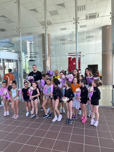 In Sancaktepe, Turkey, a group of young girls, dressed in leotards and holding bouquets of flowers, stand outside a modern building with large glass doors and windows. A man and two women, presumably coaches or chaperones, stand with the girls. The group is gathered on a tiled walkway in front of the building. Behind them, a Turkish flag is visible hanging on the wall. The overall atmosphere appears to be celebratory, possibly after a performance or competition.