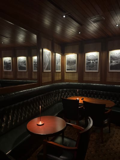 The image presents an unoccupied interior space, identified as a lounge or bar located in Sankt Moritz, Switzerland. The room is characterized by dark wood paneling on both the walls and ceiling, with several recessed spotlights providing ambient illumination. An L-shaped banquette, upholstered in dark green tufted leather, lines the perimeter. Multiple round wooden tables are positioned in front of the banquette, each featuring a slender, copper-toned table lamp emitting an orange glow. Dark wooden chairs with upholstered seats are arranged around the tables. The walls display numerous framed black and white landscape photographs, primarily depicting mountains and lakes, each illuminated by an individual brass picture light. A large mirror on the left wall reflects portions of the room and additional artwork. The floor is covered with a dark, patterned carpet. No individuals are present in the scene.